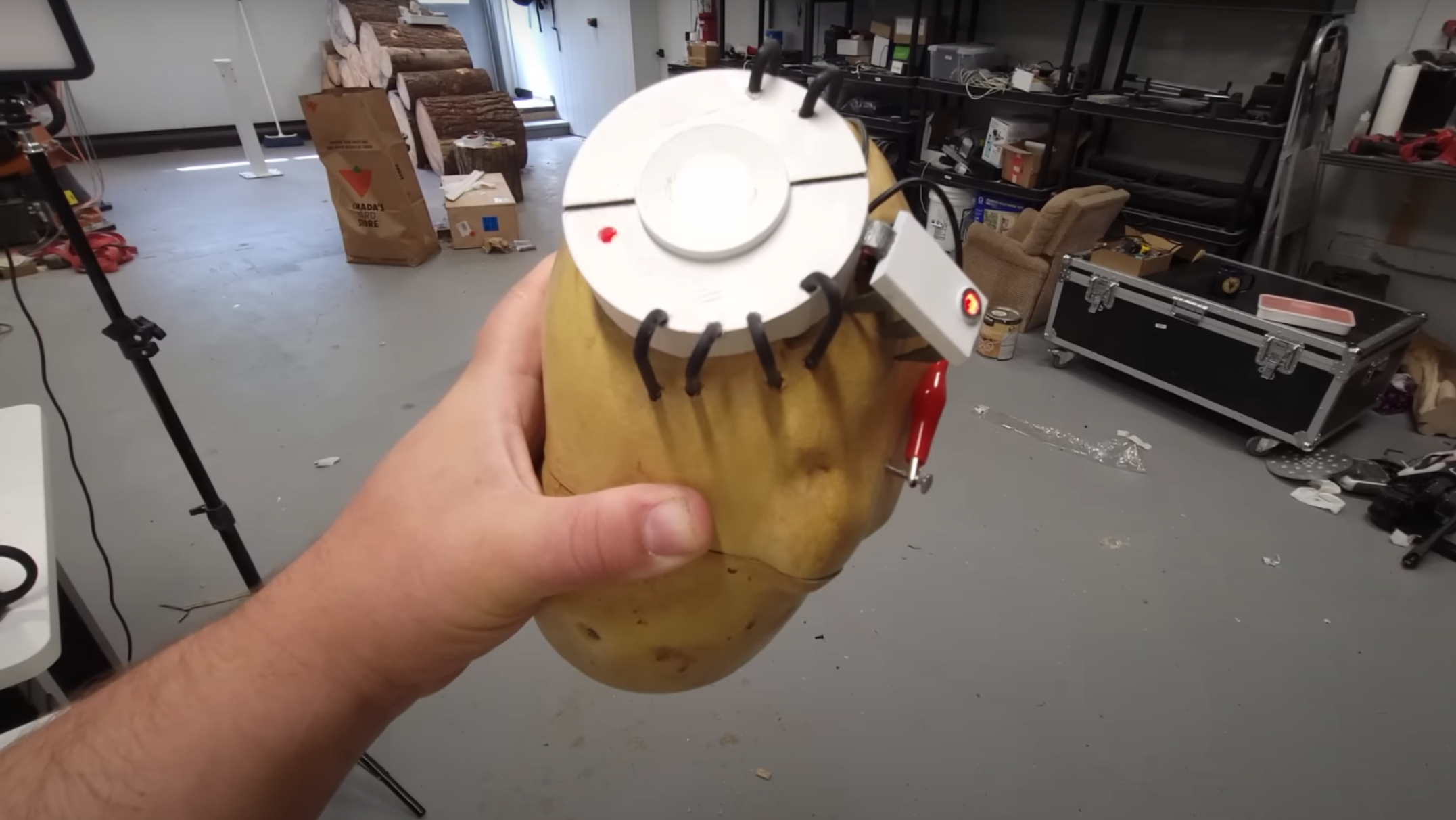 A man’s hand is visible holding a large, potato-shaped object in the foreground. A short, white, cylindrical structure is on the top of the potato, with black wires bending back into the potato. A smaller rectangular structure is to one side of it, and a red alligator clip connects to a nail protruding from the potato.