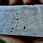 A man’s hand is shown holding a polished metal billet. The billet has a few voids in the surface, and the surface shows a pattern of lighter lines against the darker metal background.